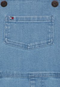 Lichtblauwe denimstof heeft een achterzak met een rechthoekige vorm en zichtbare stiksels. Een klein merkje is aanwezig op de zak.