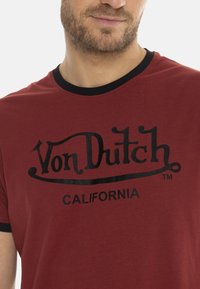 T-shirt en coton rouge avec un col rond noir et des manches courtes. Arbore un logo "Von Dutch" en noir audacieux et le texte "CALIFORNIA" en dessous.
