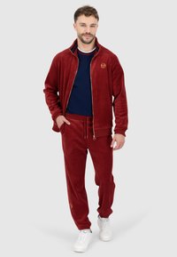 Roter Samt-Trainingsanzug mit einer Jacke mit Reißverschluss und einem goldenen Logo. Dazu passende Hose und weiße Turnschuhe. Glatte Textur.