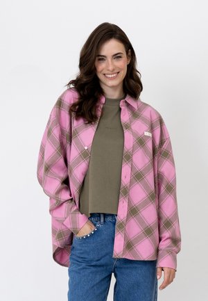 Ung kvinde med langt brunt hår smiler iført en pink og brun ternet skjorte over en olivengrøn top og blå jeans med juveldekoration på lommerne.