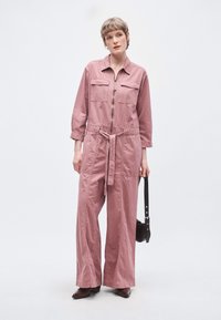 Rózsaszín farmer overál hosszú ujjakkal, két mellzsebbel, öves derékrésszel, bő szárakkal és elülső cipzárral. A modell egy kis fekete táskát tart.