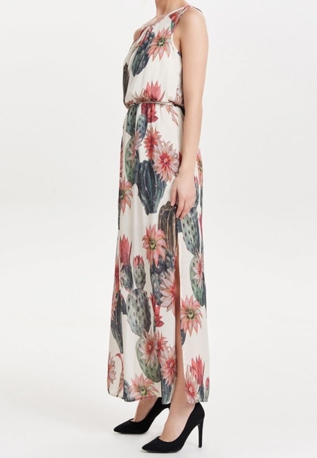 Femme portant une robe maxi blanche sans manches avec un grand imprimé de fleur de cactus rose et verte, et des talons noirs, debout de profil.