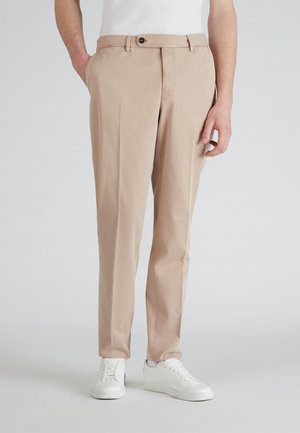 Beige katoenen broeken met een vlakke voorkant, zijzakken en een enkele knoopsluiting. Draagt witte sneakers tegen een lichte achtergrond.