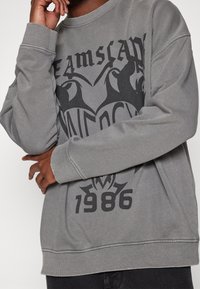 Sudadera gris con un gran diseño gráfico negro, puños acanalados y un escote redondo. El texto incluye "1986" en la parte inferior.