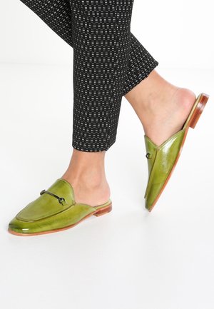 Zielone skórzane mule z ostrym noskiem, płaskim, drewnianym obcasem i ozdobnymi czarnymi okuciami. Noszone z czarnymi, wzorzystymi spodniami.