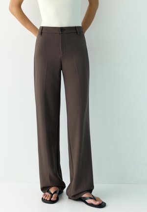 Person wearing high-waisted dark brown wide-leg trousers with front crease, paired with black flip-flops, standing against plain light background.