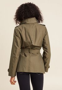 Olijfgroene jas met ceintuur, een rechtopstaande kraag, gestructureerde schouders en een gedefinieerde taille, met een achterflap en lange mouwen.