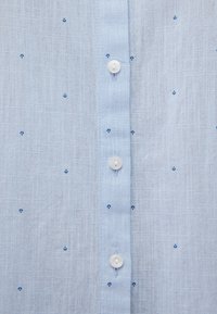 Himmelblaues Hemd mit Knöpfen aus strukturiertem Stoff, das kleine marineblaue Muster und weiße Kunststoffknöpfe entlang der Vorderblende aufweist.