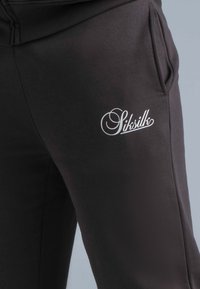 Zwarte joggers van zachte stof, met zijzakken en een wit logo op de dij. Soepele textuur met een comfortabele pasvorm.