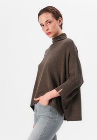 Pull en mélange de laine marron avec un col montant, des épaules tombantes et des fentes sur les côtés. Associé à un jean gris ajusté. Texture lisse et douce.