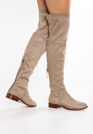 Bottes beige en daim montantes au-dessus du genou avec talons bas en bois et fermetures éclair latérales, portées par une personne debout sur un sol blanc.