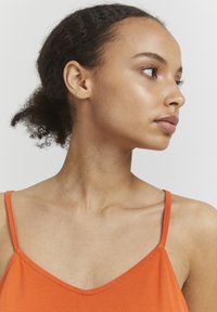 Orange ærmeløs top med v-hals design. Glat stof, tynde stropper og en tætsiddende silhuet. Hudtonen er medium med en naturlig finish.