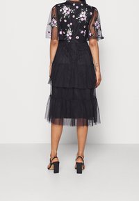 Robe en tulle noire avec un ourlet en couches, ornée d'une broderie florale sur des manches transparentes, présentant un corsage ajusté et une fermeture éclair dans le dos.