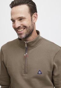 Felpa marrone con zip e colletto alto, in tessuto testurizzato, e un piccolo logo triangolare blu sul petto. La zip presenta dettagli rossi e blu.