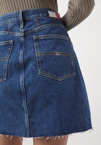 Jeanskjol i mörkblått med rå kant. Har gula sömmar och två bakfickor. Tommy Jeans-logotagg vid midjan.