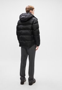 Μαύρο μπουφάν με κουκούλα, με υφή καπιτονέ και ορατές ραφές. Φορεμένο πάνω από γκρι παντελόνι και μαύρα παπούτσια.