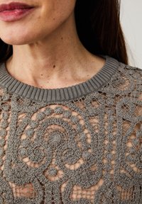 Haut gris en tricot avec un dessin en dentelle complexe comprenant des volutes et des cercles, complété par un col côtelé. Texture texturée et tissage ajouré.
