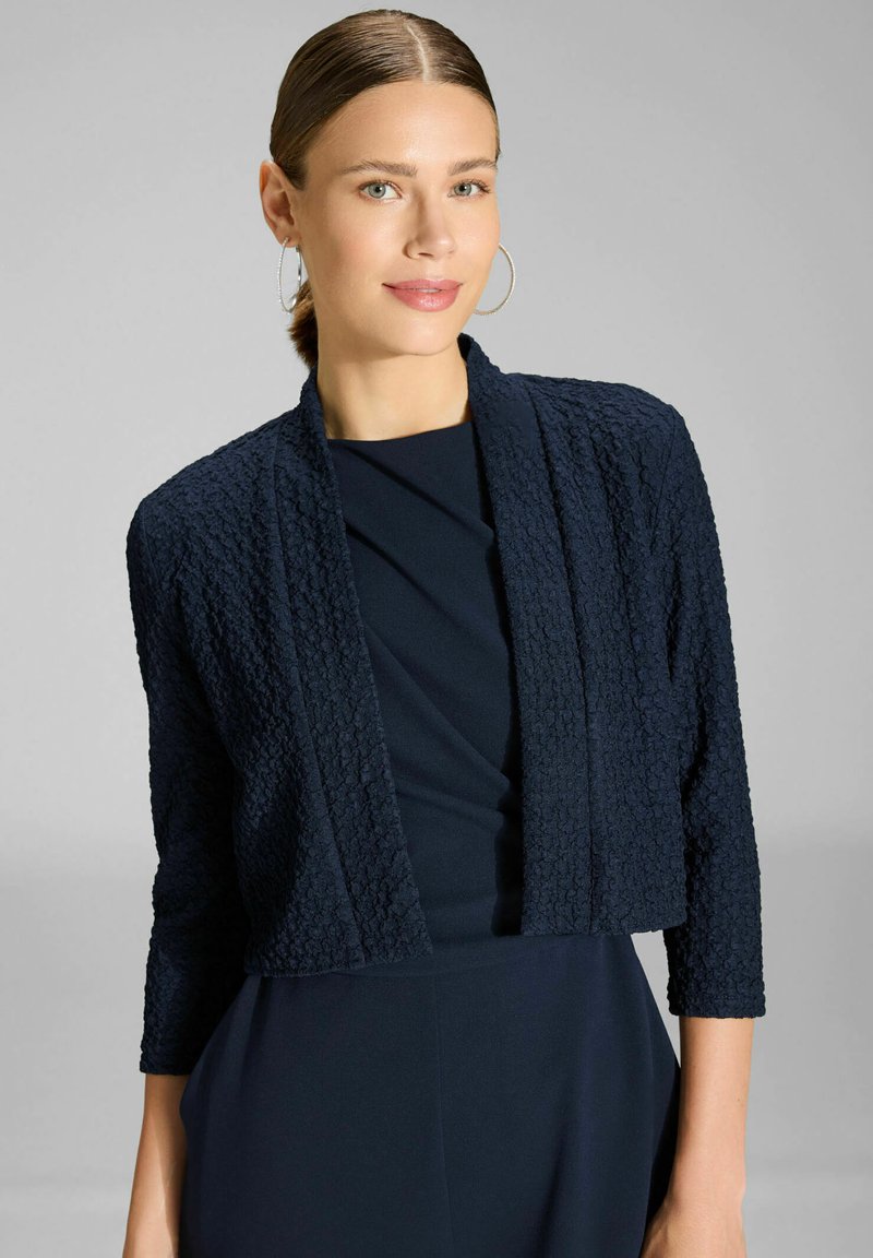 Femme aux cheveux lissés en arrière, portant de grandes boucles d'oreilles en anneau et une veste courte texturée bleu marine sur une robe bleu marine, face à l'avant, fond neutre.
