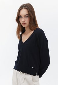 Suéter de v-neck en azul marino hecho de un material de punto suave, con ribetes en el dobladillo y aberturas laterales. Presenta un pequeño logo cerca del dobladillo.