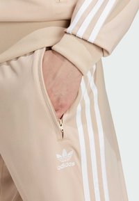 Beige träningsbyxor med vit tre-randsdesign, ribbade mudd, dragkedjeficka och broderad Adidas-logga på vänster lår.