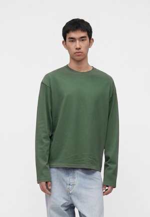 Giovane uomo con capelli corti neri che indossa una camicia verde lunga e larga e jeans azzurri, in piedi su uno sfondo bianco semplice.