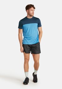 Blå och marinblå sportt-shirt med korta ärmar, kombinerad med svarta shorts, iförd grå strumpor och svarta löparskor, står avslappnat.
