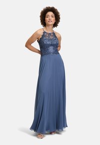 Longue robe bleue avec un corsage en dentelle et une superposition transparente. Design à col halter et jupe fluide. Complétée par de délicates bretelles et un subtil éclat.