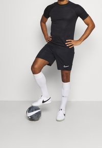 Camiseta deportiva negra y pantalones cortos, logo de Nike blanco, calcetas blancas hasta la rodilla y zapatillas deportivas negras, con un balón de fútbol negro y gris.