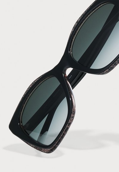 Czarne okulary przeciwsłoneczne o dużych, kwadratowych oprawkach. Krawędzie mają metaliczny akcent o teksturowanej powierzchni, a soczewki są ciemnoszare.