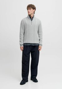 Hellgrauer Strickpullover mit hohem Kragen, ausgestattet mit einem Reißverschluss, Zopfmuster und gerippten Bündchen, kombiniert mit dunklen Hosen und Schuhen.
