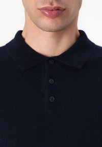 Navy modrý pletený polo tričko s jemnou texturou, které má límec a čtyři tmavé knoflíky podél předního zapínání.