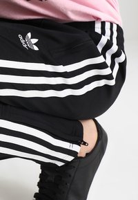 Czarne sportowe spodnie dresowe z białymi paskami, z logo Adidas i zamykaną kieszenią na zamek. Do tego czarne sznurowane trampki.