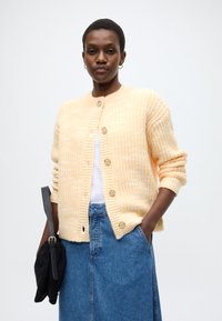 Κίτρινη πλεκτή ζακέτα με ριμπ υφή, στρογγυλή λαιμόκοψη και πέντε κουμπιά. Συνδυασμένη με μια φούστα τζιν και κρατώντας μια μαύρη τσάντα.