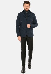 Giacca trapuntata blu scuro con colletto, bottoni frontali, due tasche con zip e un motivo testurizzato. Indossata sopra un top nero a collo alto e pantaloni.