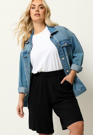 Blondine vrouw draagt een wit T-shirt, een blauwe spijkerjas met opgerolde mouwen en losse zwarte korte broek met hand in de zak.