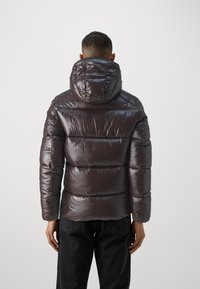Glimmende donkerbruine pufferjas met capuchon, gestikte secties en een getailleerd silhouet. Draagt over donkere jeans, gezien vanaf de achterkant.