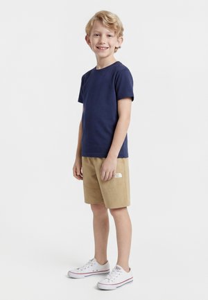 Niño pequeño sonriente, vistiendo camiseta azul marino, pantalones cortos beige con logo y zapatillas blancas, sobre un fondo blanco liso.