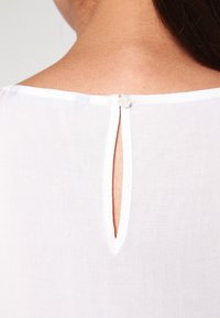 Haut en tissu blanc léger avec un col rond et une petite ouverture en forme de goutte d'eau à l'arrière, fermée par un bouton. Texture lisse.