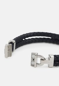 Fekete fonott bőrszíj ezüstszínű csattal, amelyen gravírozott "HILFIGER" felirat és kör alakú díszítések találhatók. Texturált felület és letisztult dizájn.