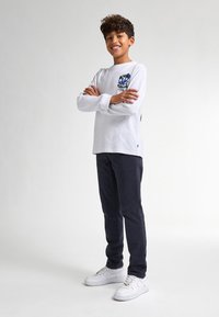 Camicia bianca a maniche lunghe con dettagli grafici blu, abbinata a pantaloni scuri e sneaker bianche. Design a taglio dritto, materiale cotone.