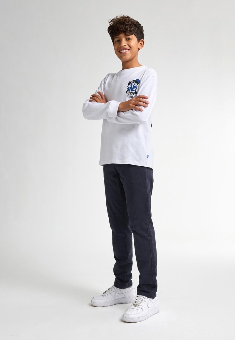 Camicia bianca a maniche lunghe con dettagli grafici blu, abbinata a pantaloni scuri e sneaker bianche. Design a taglio dritto, materiale cotone.
