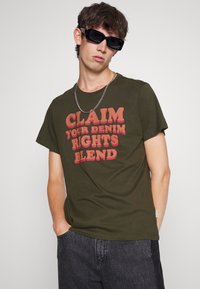 Olivenfarsket bomulds T-shirt med fed rød teksttryk, der står "HEV DINE DENIMRETTIGHEDER BLAND," med en afslappet pasform og korte ærmer.