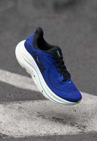 Modré a černé závodní boty Hoka s tlustou bílou podrážkou ve vzduchu nad asfaltovou silnicí se bílými silničními značkami.