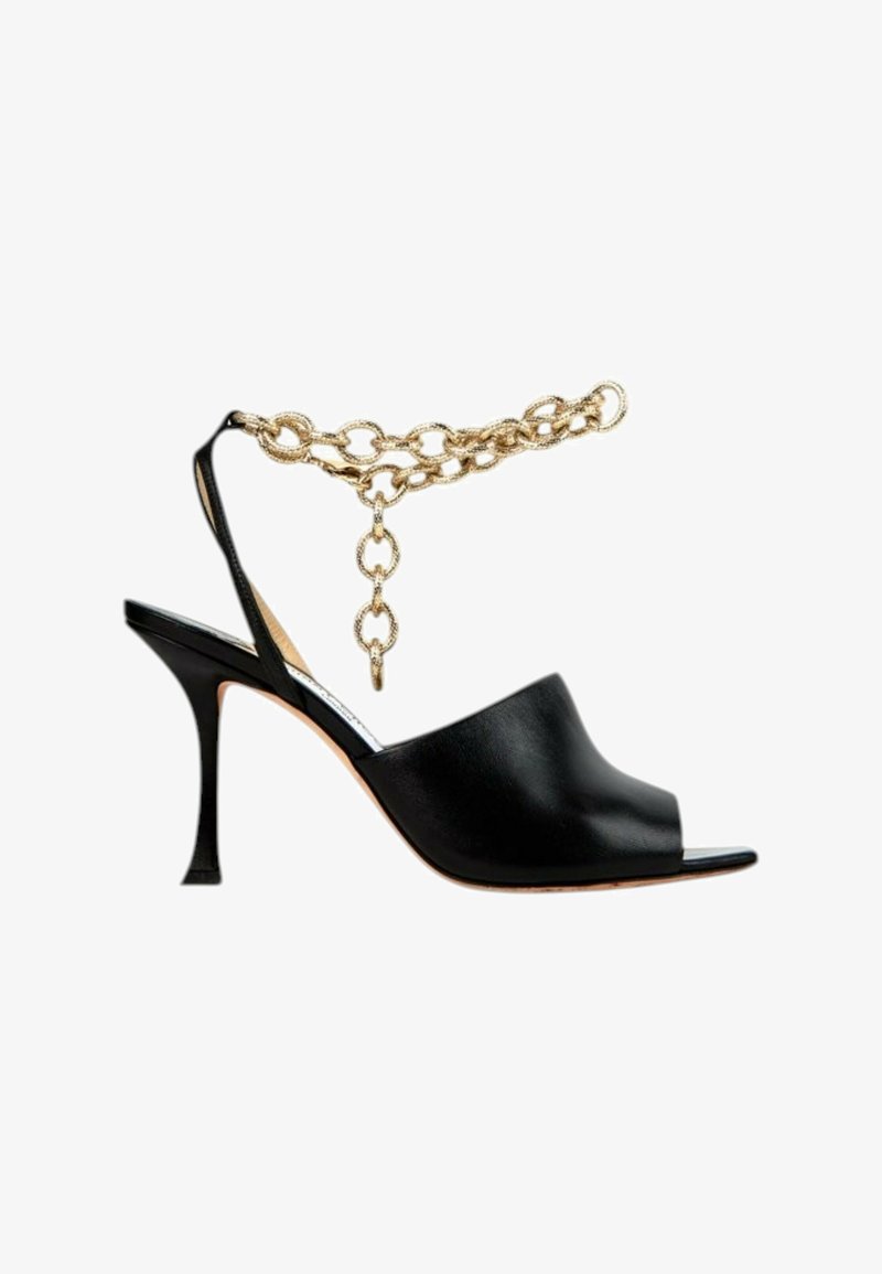 Chaussure noire à talon haut et bout ouvert avec bride arrière et décoration chaîne dorée à la cheville sur fond blanc.