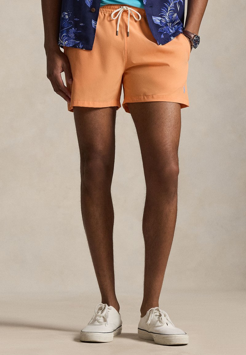 Άνδρας που φοράει πορτοκαλί σόρτς με κορδόνι, λευκά αθλητικά παπούτσια και μπλε πουκάμισο με λουλούδια, με το ένα χέρι στην τσέπη, μπροστά σε μπεζ φόντο.