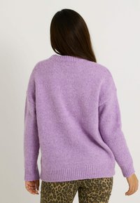 Persona con cabello largo y oscuro, vistiendo un suéter de punto lavanda y pantalones de leopardo, vista desde atrás contra un fondo simple.