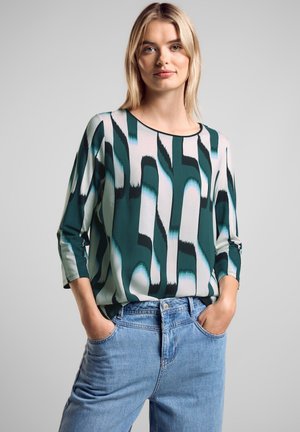 Blusa verde e bianca con motivo astratto, scollatura rotonda e maniche a tre quarti, abbinata a jeans in denim azzurro chiaro.