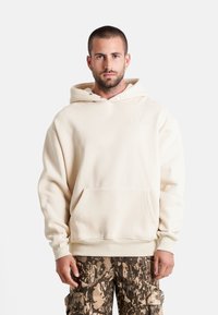 Sudadera color crema hecha de un tejido suave, con un bolsillo delantero, capucha con cordón y un detalle de logo minimalista. Combinada con pantalones de camuflaje.