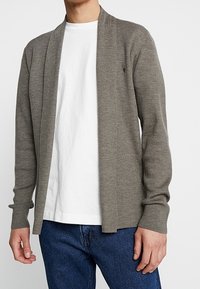 Grey, ribbed cardigan with an open front design. Features long sleeves and a subtle logo on the chest. Worn over a white t-shirt.