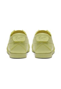 Keltainen nahkakengät, joissa on kumipohjat, joissa on ommeltu yksityiskohtia ja logon päällyste kantapäässä. Minimalistinen muotoilu ja pyöristetty muoto.